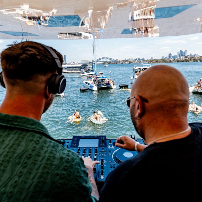 Two DJs are performing on a boat with a city skyline and bridge in the background. They're facing a group of anchored boats and people swimming. The scene is lively and sunlit.