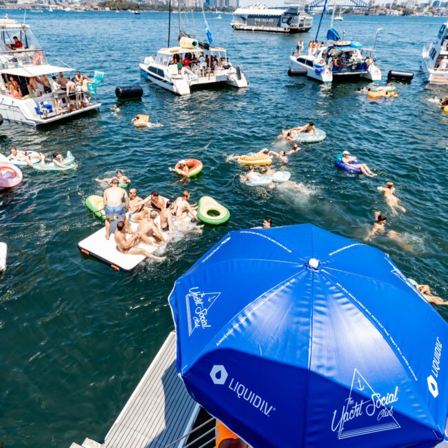 People relaxing on floats in a sunny bay near several boats. A large blue umbrella is in the foreground with city skyscrapers and a bridge visible in the background. Steps lead down to the water.