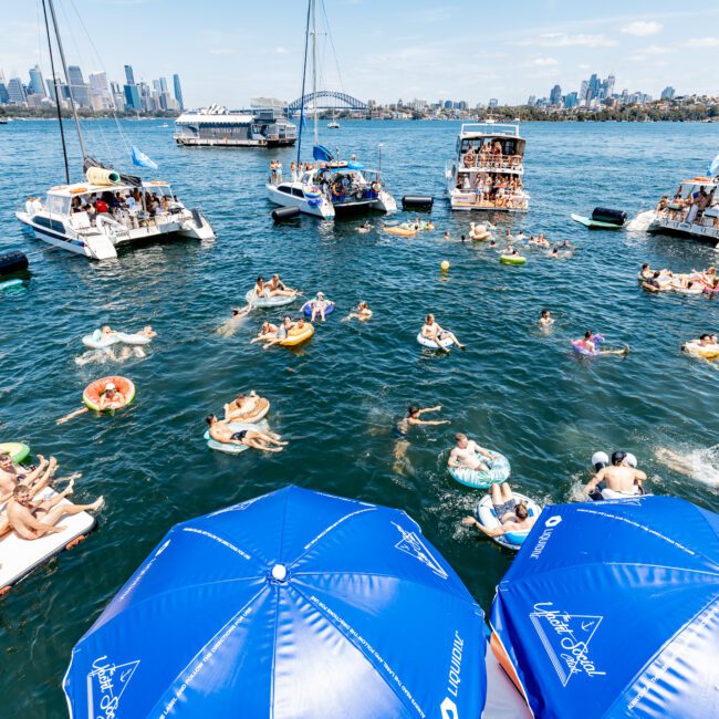 Groups of people relax on boats and float in the water near a city skyline. Many are on inflatable rafts, with blue umbrellas in the foreground. It's a sunny day, and the scene looks lively and festive.