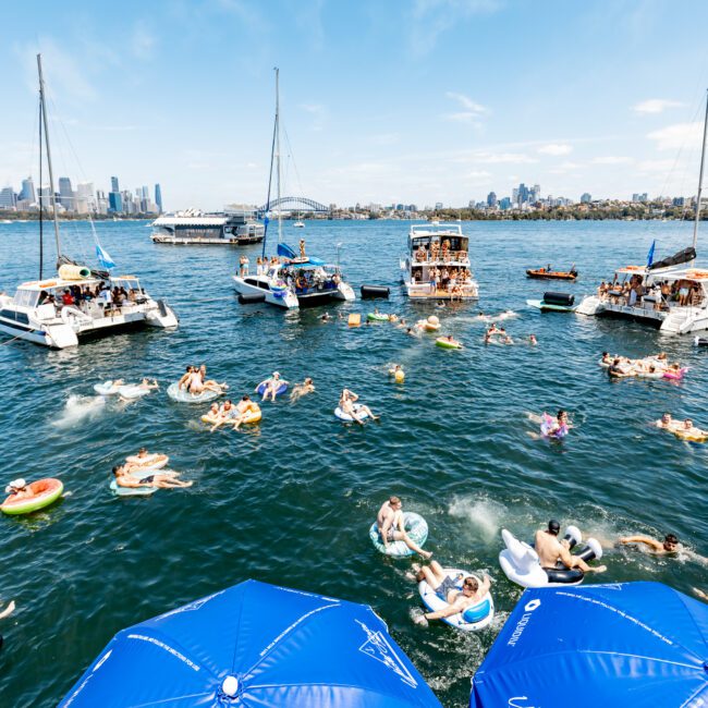 A busy scene on a sunny day with numerous people floating on inflatable tubes in a lake. Several boats are anchored nearby while the city skyline and a bridge are visible in the distance. Blue umbrellas are in the foreground.