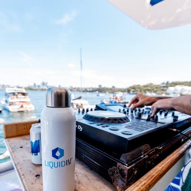 A Liquid I.V. bottle stands on a table beside DJ equipment on a boat. In the background, multiple boats are visible on the water under a blue sky.