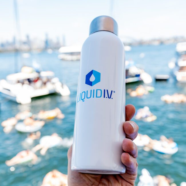 A hand holds a white Liquid I.V. bottle against a blurred background of a lively scene with people swimming and floating on inflatable rafts in a sunny body of water with boats around.