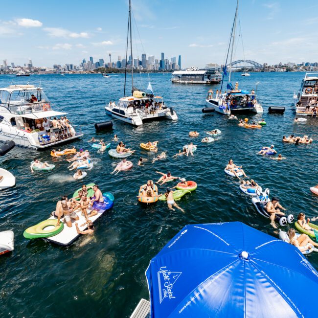 A vibrant scene of people enjoying a sunny day on a bay. Groups relax on inflatable floaties between anchored boats. The city skyline and a bridge are visible in the background. A blue umbrella with text is in the foreground.