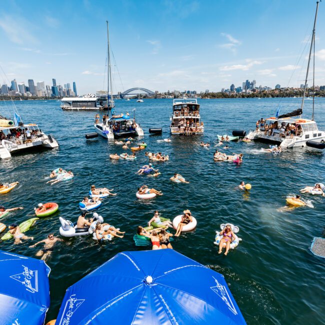 People are enjoying a sunny day on a lake, swimming and floating on inflatables. Several boats are anchored nearby, and a city skyline is visible in the background. Blue umbrellas are in the foreground, providing some shade.