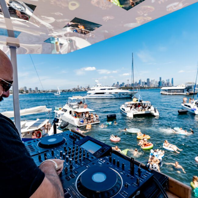 A DJ wearing headphones is performing on a boat deck with turntables. The background shows several boats and people on inflatables enjoying the water. A city skyline with tall buildings is visible under a clear blue sky.