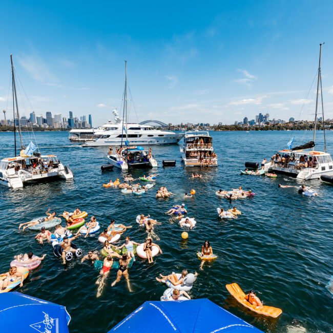 A lively gathering on a sunny day with people swimming and floating on inflatables between anchored boats. The city skyline is visible in the background under a clear blue sky. Sailboats and a large yacht are also present.