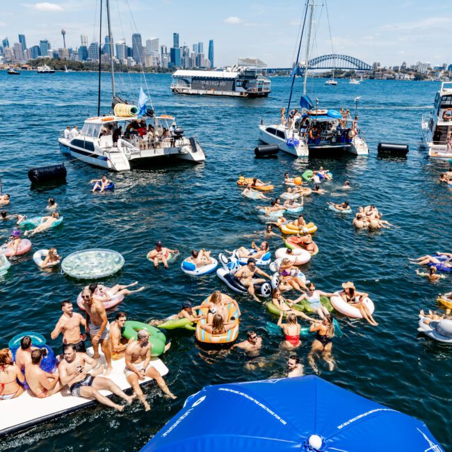 A lively scene of people in swimsuits enjoy floating on inflatable devices in a vibrant blue harbor. Boats are anchored nearby, with a city skyline and bridge in the background. A blue umbrella is in the foreground.