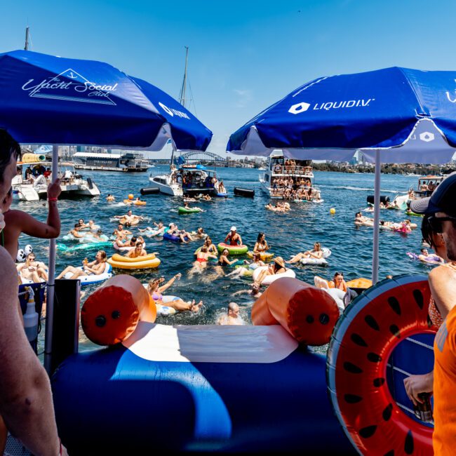 People enjoying a sunny day on a lake surrounded by boats. Many are on inflatables near blue umbrellas labeled "Liquid IV" and "The Yacht Social Club." The scene is lively and festive.