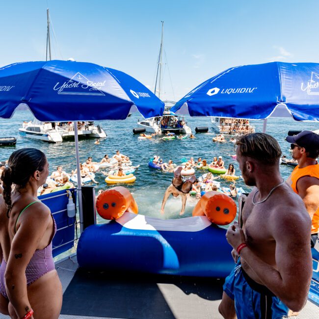 People in swimwear gathered on a boat deck with blue umbrellas and others in the water on inflatable floats. Boats are anchored in the background on a sunny day.