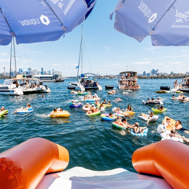 A lively gathering of people on colorful inflatables in the water surrounded by boats. Umbrellas provide shade, while a distant city skyline is visible under a clear blue sky.