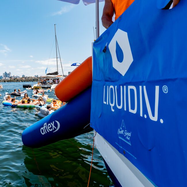 A group of people relax on inflatable rings and pool floats in the water near a boat with a "LIQUID IV" banner. Other boats are visible in the background under a clear blue sky.