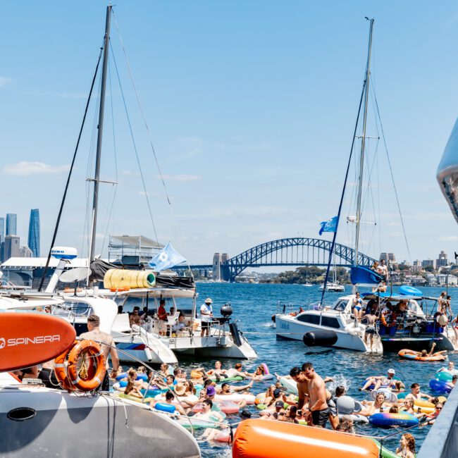 Boats and people on inflatables gather on a sunny day in a harbor. A prominent bridge is visible in the background. The scene is lively, with clear blue skies and numerous colorful floats in the water.