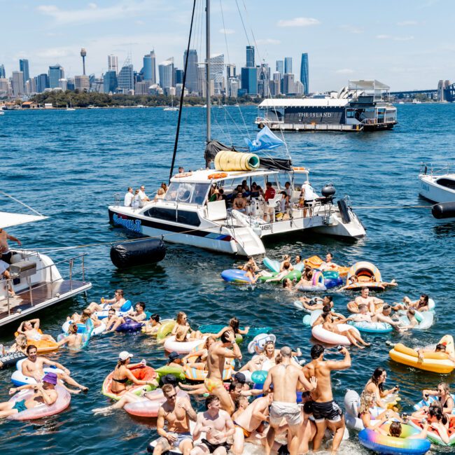 A lively scene on the water with a large group of people enjoying a sunny day, floating on inflatable pool toys near a boat. The city skyline is visible in the background under a clear blue sky.