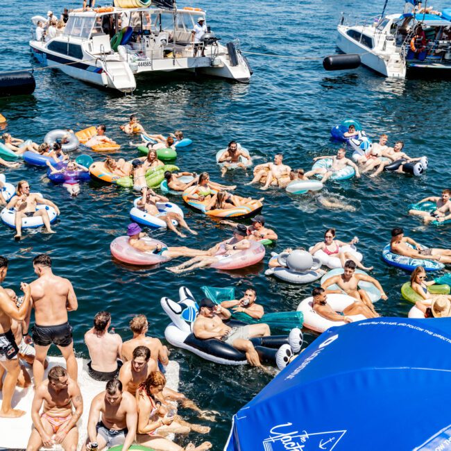 People enjoying a sunny day on floating inflatables and boats in a body of water. The city skyline is visible in the background. There is a blue umbrella with "Uptown Social" written on it in the foreground.