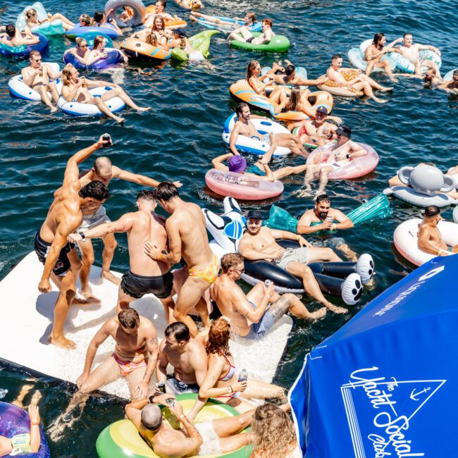 A lively group of people enjoying a sunny day on the water with inflatables and a boat nearby. Some are sunbathing, others are swimming, and colorful floats are scattered around. A blue umbrella reads "Yacht Social Club.