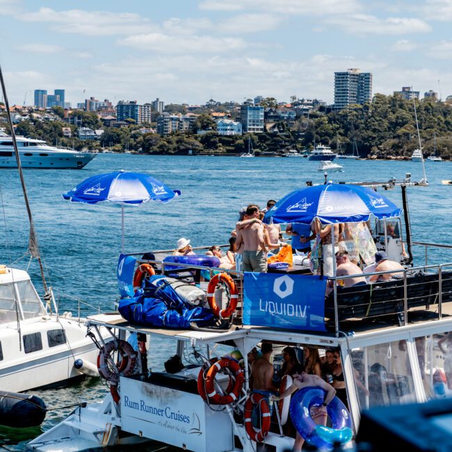 A lively scene on a boat with people gathered on the deck, some under blue umbrellas, enjoying the sunny day. The boat is named "Rum Runner Cruises" and is on a picturesque body of water with a city skyline in the background.