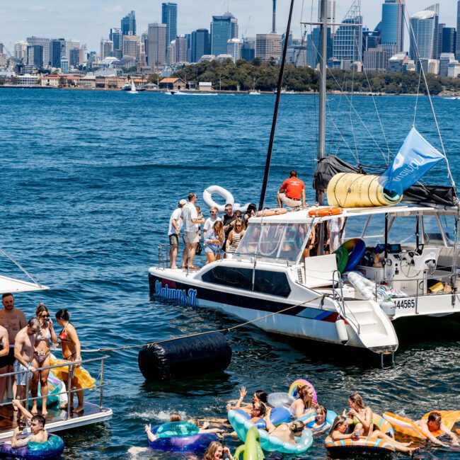 People enjoy a sunny day on the water, floating on inflatables near a boat. The city skyline is visible in the background. The atmosphere is lively and cheerful.