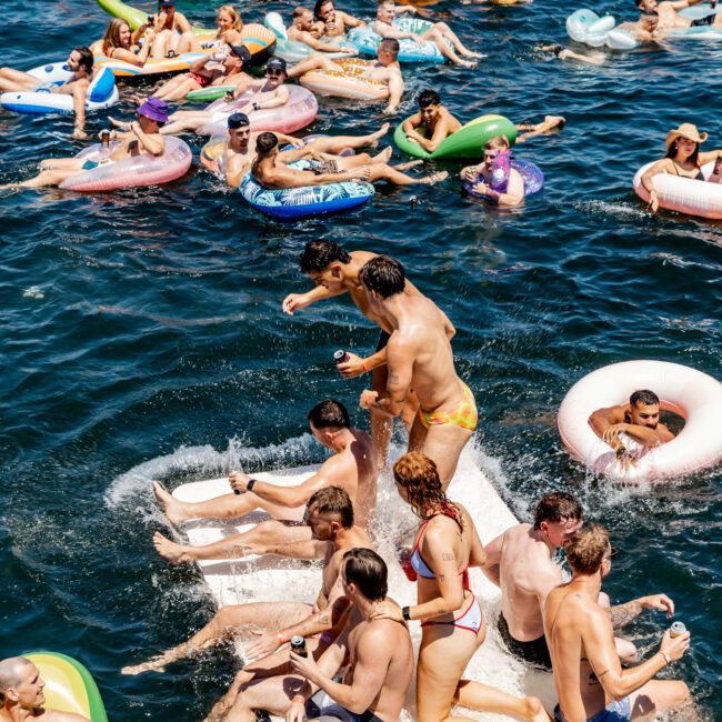 A large group of people enjoying a sunny day on the water. They are floating on inflatable rings and rafts, with some relaxing and others socializing. A boat is visible in the background. The atmosphere is lively and festive.