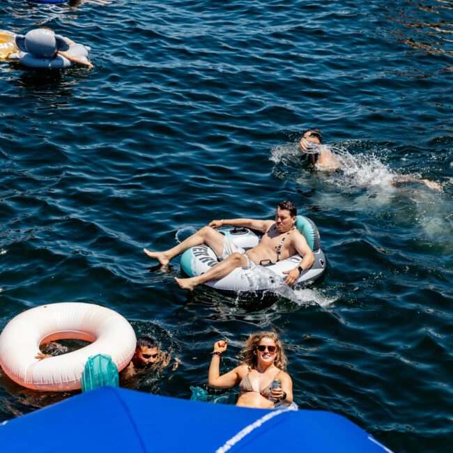 People enjoying a sunny day on a lake, lounging on inflatable floats. Others swim nearby. A large blue umbrella with text partially visible is in the foreground.