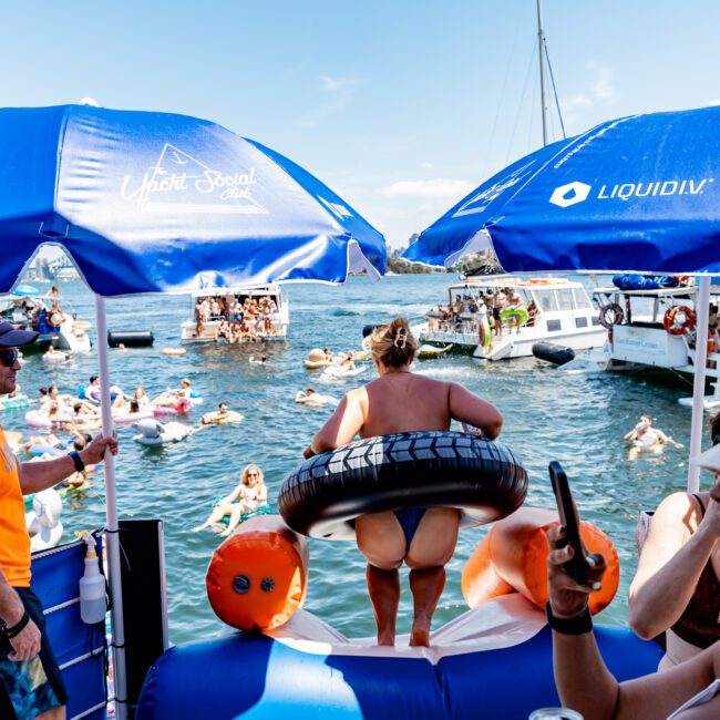 People enjoying a sunny day on boats and inflatables in the water. Blue umbrellas with "Liquid I.V." branding provide shade. One person holds an inner tube, while others relax with drinks and take photos. Boats are filled with more people in the background.