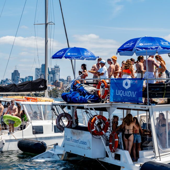 A group of people enjoying a sunny day on a party boat, with umbrellas labeled "LIQUID" on the upper deck. The boat is named Rum Runner Cruises. The city skyline is visible in the background. People are wearing swimsuits and hats.