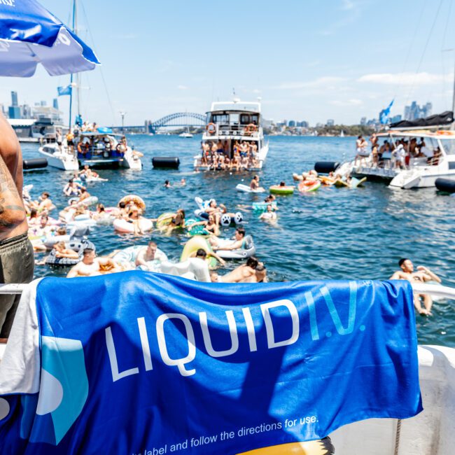 People are enjoying a sunny day on the water near boats. A blue Liquid IV towel hangs prominently in the foreground. In the background, individuals are on inflatable rafts and swimming, with a city skyline visible.