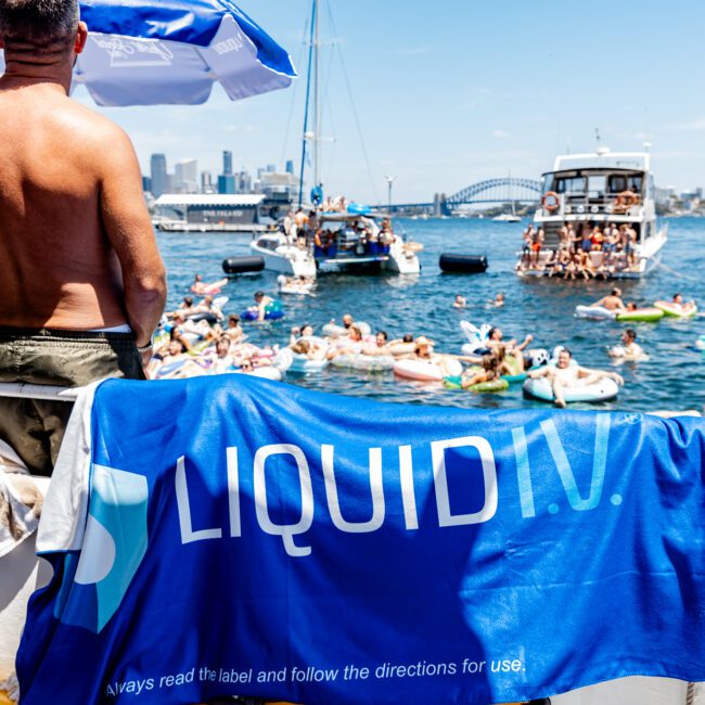 A lively scene on a sunny day with people swimming and floating on inflatables in the ocean near several boats. A large blue towel with "LIQUID I.V." hangs over a railing. The skyline is visible in the background under a clear blue sky.