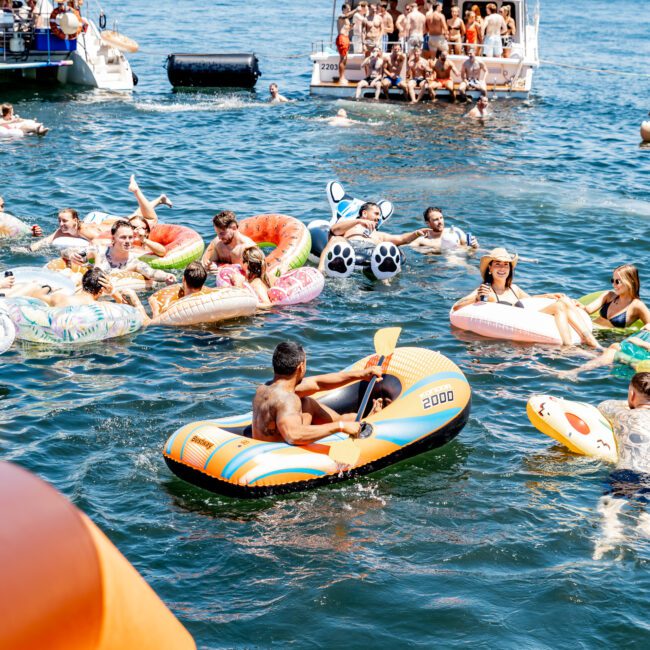 People on inflatable boats and pool floats enjoying a sunny day on the water. A yacht is in the background, along with a bridge and city skyline. The atmosphere is lively and festive.