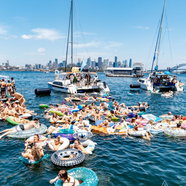 People are floating on inflatable toys in a sunny bay with city skyscrapers in the background. Two boats are anchored nearby. The scene is lively with participants enjoying the water and sun.