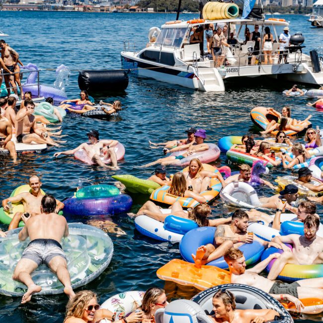 A lively scene with people enjoying a sunny day on a lake, floating on colorful inflatables. A boat with a water cannon is anchored nearby, and a city skyline is visible in the background.