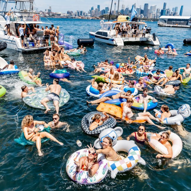 A lively scene with people floating on inflatable tubes and loungers in a body of water near anchored boats. The city skyline is visible in the background. Participants are enjoying a sunny day, socializing, and relaxing.