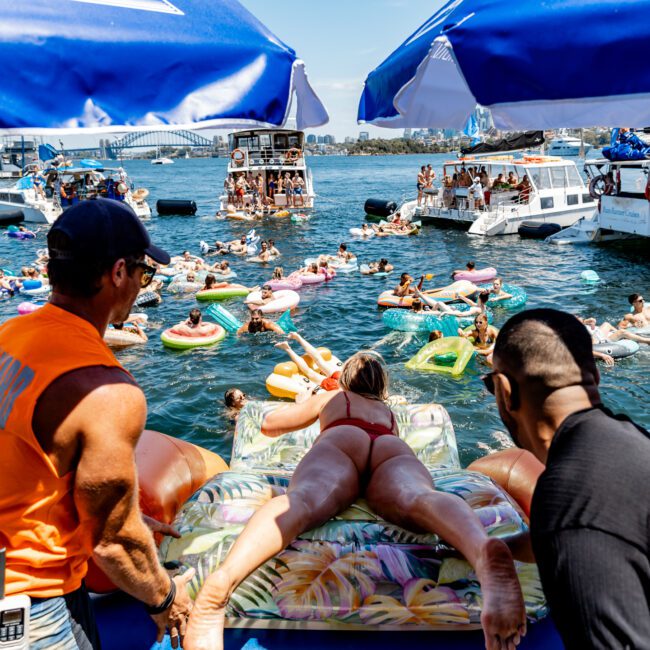 People enjoying a boat party with many lounging on inflatables in the water. Two people on a boat are pushing a woman lying on a large inflatable raft. The scene is lively with several boats and a bridge visible in the background.