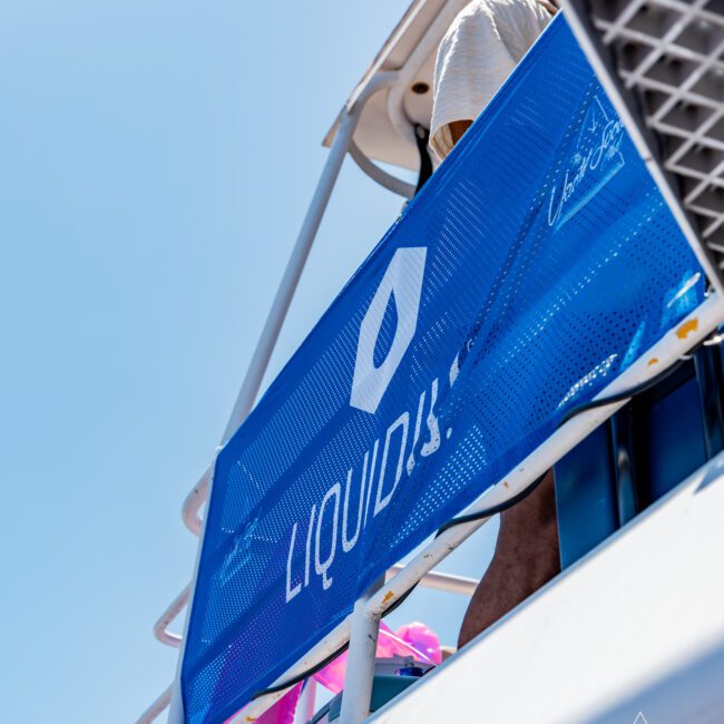 Close-up of a blue banner with the word "LIQUID" on a boat railing. The sky is clear and blue, and part of the boat structure is visible. The logo and text "The Yacht Social Club" appear in the bottom corner.