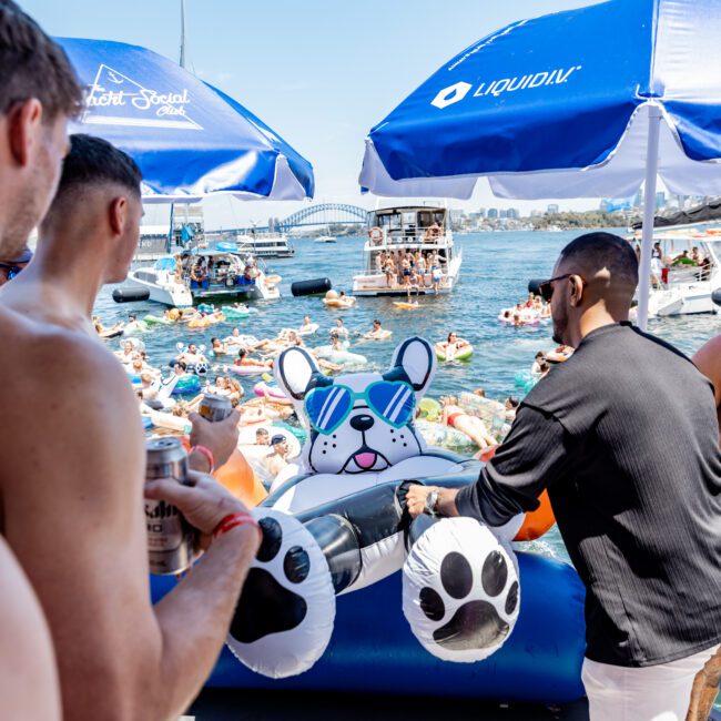 A group of people on a dockside party with inflatable pool toys. The main inflatable features a cartoon dog wearing sunglasses. There are blue umbrellas, a scenic body of water, and a boat in the background.