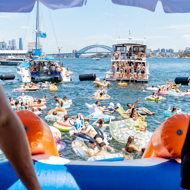 A lively scene of people on inflatable floats in the water, surrounded by boats. The Sydney Harbour Bridge is visible in the background under a clear blue sky. Beach umbrellas and a sunlit harbor create a festive atmosphere.