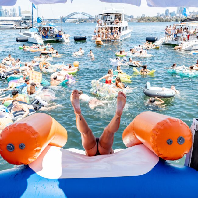 A person sliding down an inflatable orange and white slide into water, feet-first. The water is crowded with people on inflatable rings and boats. A larger boat and a city skyline, including a bridge, can be seen in the background.