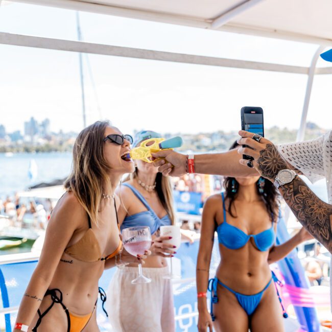 Three women are on a boat, with sunny weather. One woman in a tan bikini leans forward while another person uses a yellow toy water gun aimed towards her. Two women in blue outfits are in the background. Other boats and the city skyline are visible.