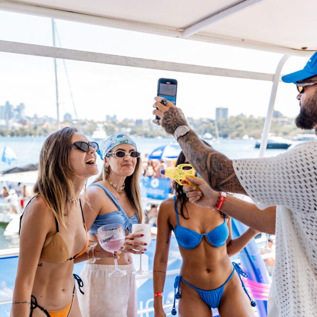 Group of people enjoying a party on a yacht. Two women in bikinis hold drinks, while a man in a hat and sunglasses takes a photo with his phone. The background shows water and distant buildings.