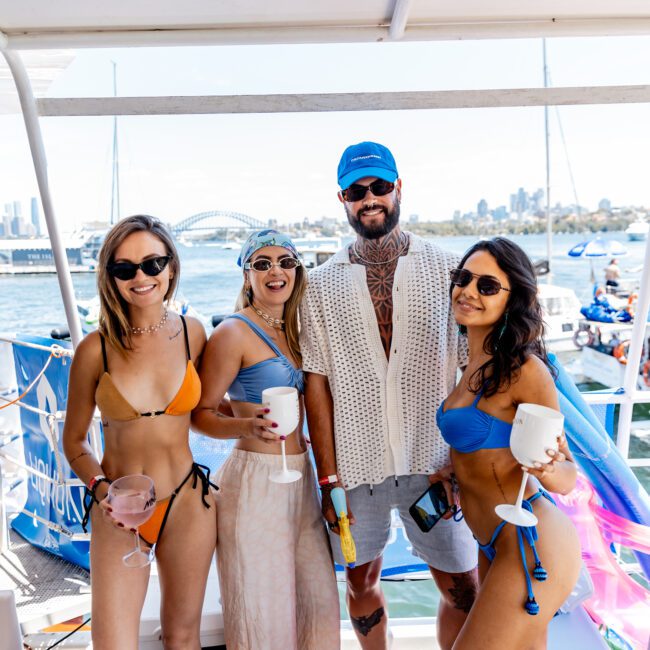 Four people posing on a boat with drinks, wearing swimwear and casual summer attire. The background shows a waterway with boats and a cityscape, including a bridge. It's a sunny day, and the group appears relaxed and happy.