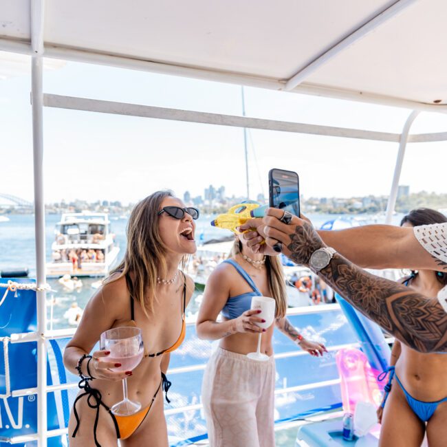 A woman in an orange bikini laughs while holding a drink on a boat. Another woman in a black bikini holds a phone and a yellow water gun. Boats and a city skyline can be seen in the background under a clear blue sky.