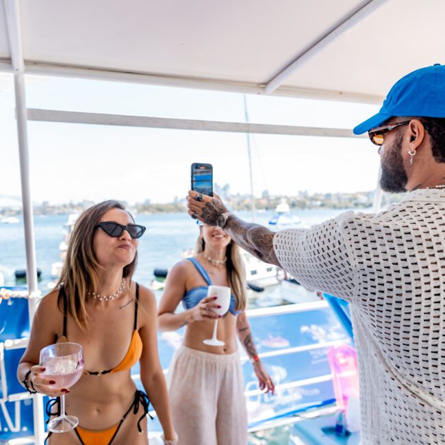 A man in a blue cap takes a photo of two women on a boat. One woman is in a bikini holding a drink, while the other wears a bikini top and light pants. They are enjoying a sunny day with water and boats in the background.