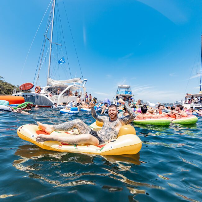 A person in patterned clothing lounges on a yellow inflatable in the water, smiling. Around them are many colorful inflatables with people, and yachts in the background, all under a clear blue sky.