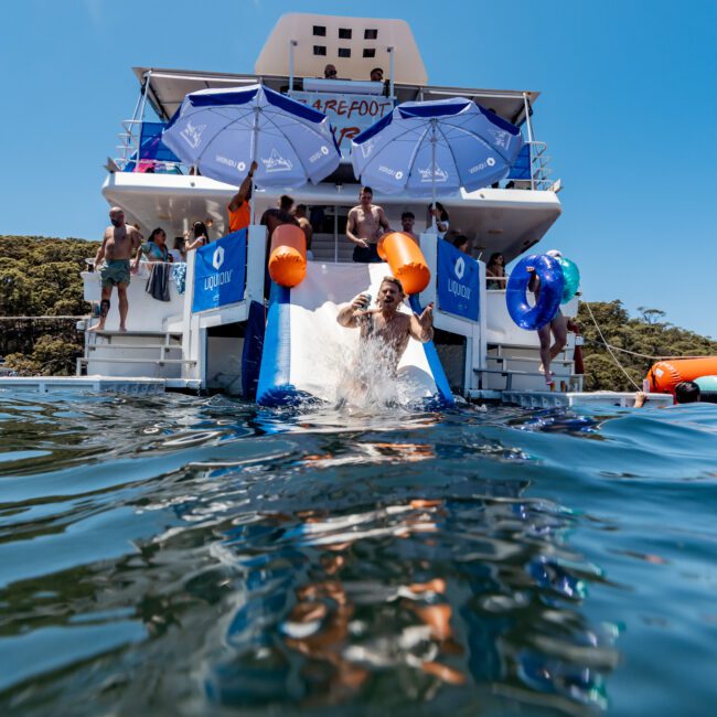 A group of people enjoys a sunny day on a yacht. Some are using a slide to splash into the water while others relax under blue umbrellas. The yacht is anchored near a green wooded shoreline.