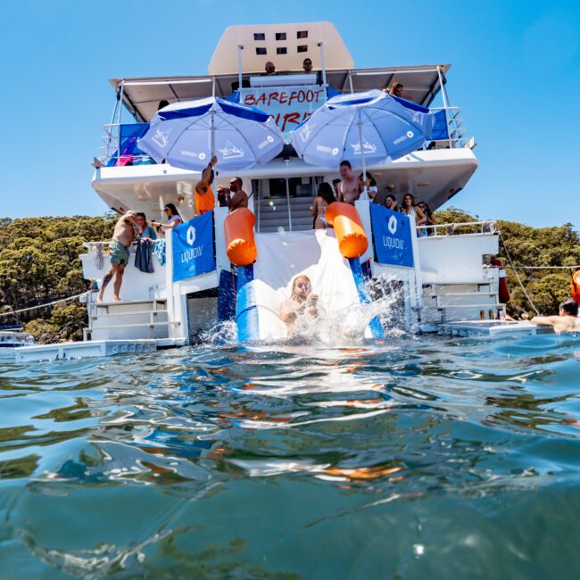 A group of people enjoy a sunny day on a yacht named "Barefoot Blue." Two people are sliding into the water from a slide on the yacht. The water in the foreground is clear, and the sky is bright and blue.