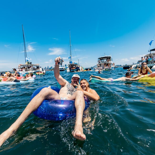 Two people joyfully float on inflatable tubes in clear blue water, surrounded by others on various floatation devices. Boats with partygoers are anchored nearby under a bright, sunny sky.