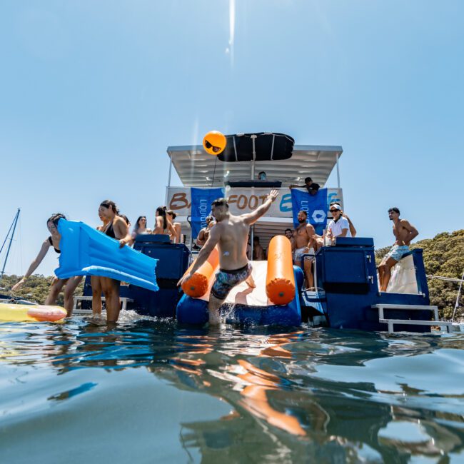 People enjoying a sunny day on a boat, sliding into the water using inflatable slides. The boat is stationary, surrounded by clear blue waters. A woman and man are on inflatables, and others are in the water. Hills are visible in the background.