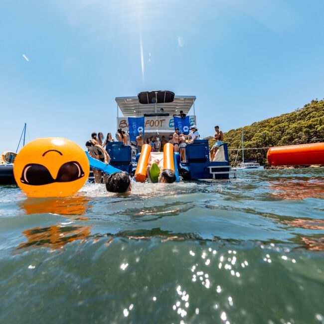 People enjoying a sunny day on a boat, with a large floating yellow emoji face in the water. Others swim and relax nearby, with lush greenery in the background.