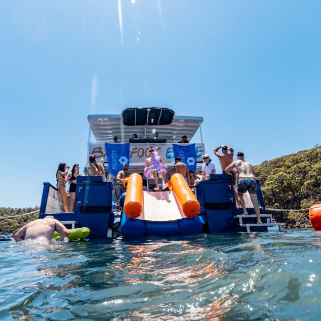 A lively scene on a yacht with people enjoying music, some dancing, others watching. A water slide leads to the ocean, and a person is swimming nearby. The sky is clear and the surrounding shores are lush with greenery.