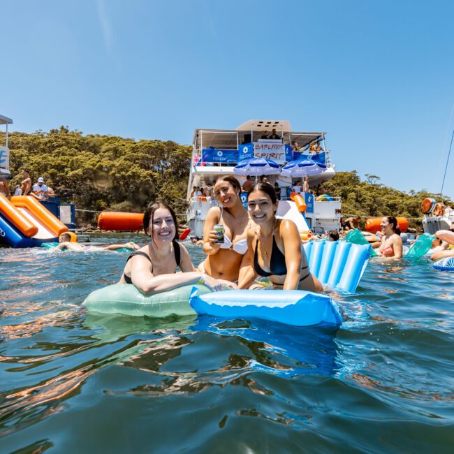 Three people smile on pool floats in a lively water setting with boats and trees in the background. Other people enjoy the sunny day in the water and on boats, with a blue sky above.