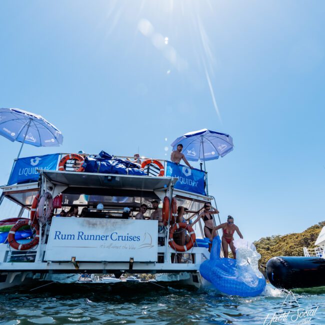 A boat named "Rum Runner Cruises" is anchored on a sunny day. People are enjoying themselves on and around the boat, with some inflatables in the water. The sun shines brightly in the clear blue sky.